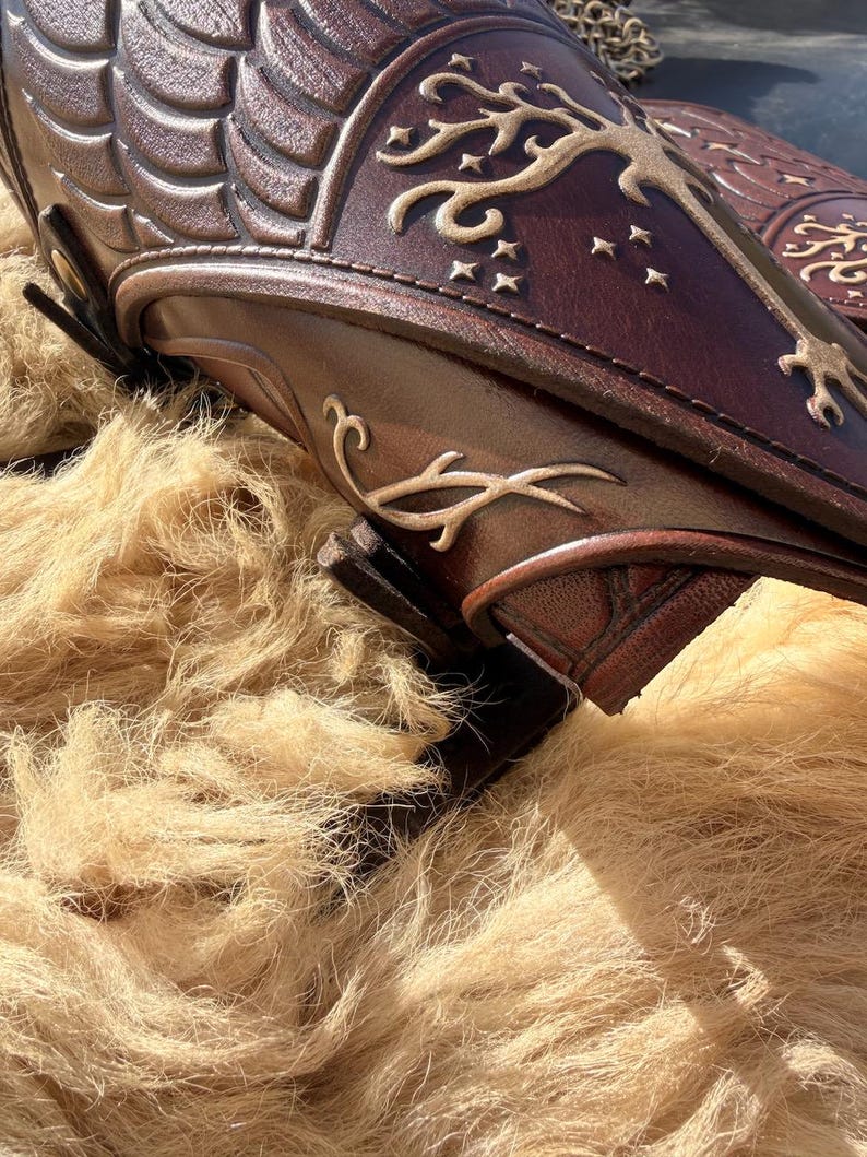 Puede incluir: Primer plano de protectores de brazo de cuero marr&oacute;n con dise&ntilde;os en relieve. Los protectores de brazo presentan patrones intrincados y un motivo de &aacute;rbol y estrellas de color dorado. El cuero tiene un color marr&oacute;n oscuro intenso con acentos marrones claros.
