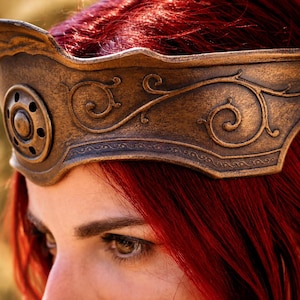 May include: A bronze-coloured headpiece featuring an embossed scrollwork pattern and a circular medallion with small perforations centred on the front. The band has a weathered, metallic finish and rests across the forehead above red hair.