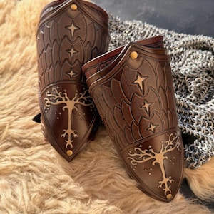 Puede incluir: Dos protectores de brazo de cuero marr&oacute;n con dise&ntilde;os en relieve de estrellas y &aacute;rboles. Los protectores de brazo tienen remaches dorados y se exhiben sobre una superficie texturizada de color marr&oacute;n claro. El dise&ntilde;o recuerda a una armadura de fantas&iacute;a.