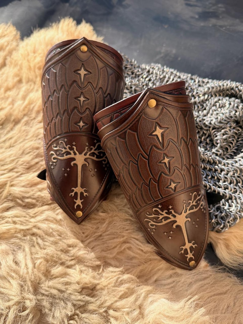 Puede incluir: Dos protectores de brazo de cuero marr&oacute;n con dise&ntilde;os en relieve de estrellas y &aacute;rboles. Los protectores de brazo tienen remaches dorados y se exhiben sobre una superficie texturizada de color marr&oacute;n claro. El dise&ntilde;o recuerda a una armadura de fantas&iacute;a.