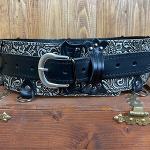 May include: A black leather belt with a silver buckle and decorative silver studs. The belt has a white floral pattern embossed on the leather.