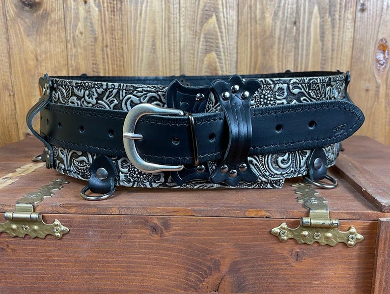 May include: A black leather belt with a silver buckle and decorative silver studs. The belt has a white floral pattern embossed on the leather.