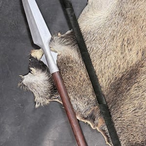 Könnte beinhalten: Ein Speer mit silberner Klinge und braunem Holzschaft. Neben dem Speer befindet sich eine schwarz umwickelte Stange. Die Objekte liegen auf einem braunen und grauen Tierfell.