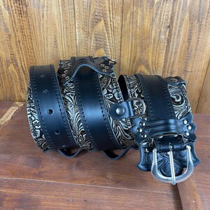 May include: A black leather belt with a silver buckle and an intricate floral design. The belt is rolled up and the buckle is visible.