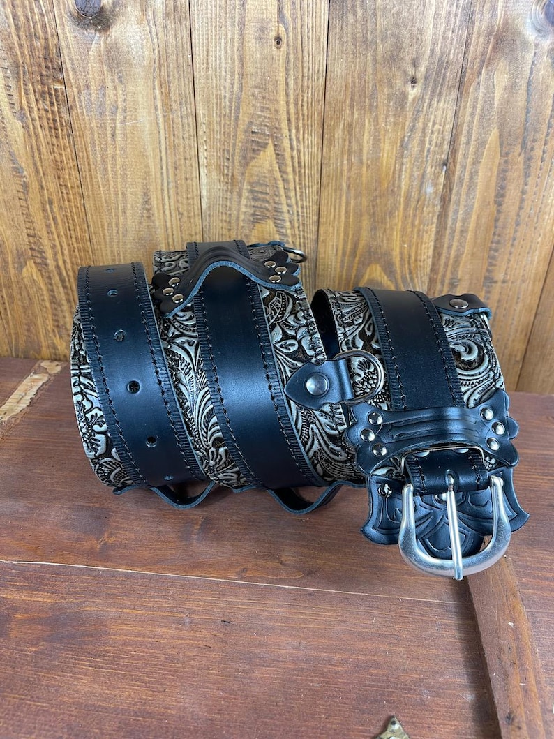 May include: A black leather belt with a silver buckle and an intricate floral design. The belt is rolled up and the buckle is visible.