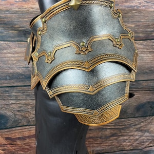 May include: A detailed, metallic shoulder armor piece in shades of gray and gold, displayed on a black mannequin. The armor features intricate embossed designs and decorative studs. The layered construction and ornate detailing suggest a fantasy or historical theme.