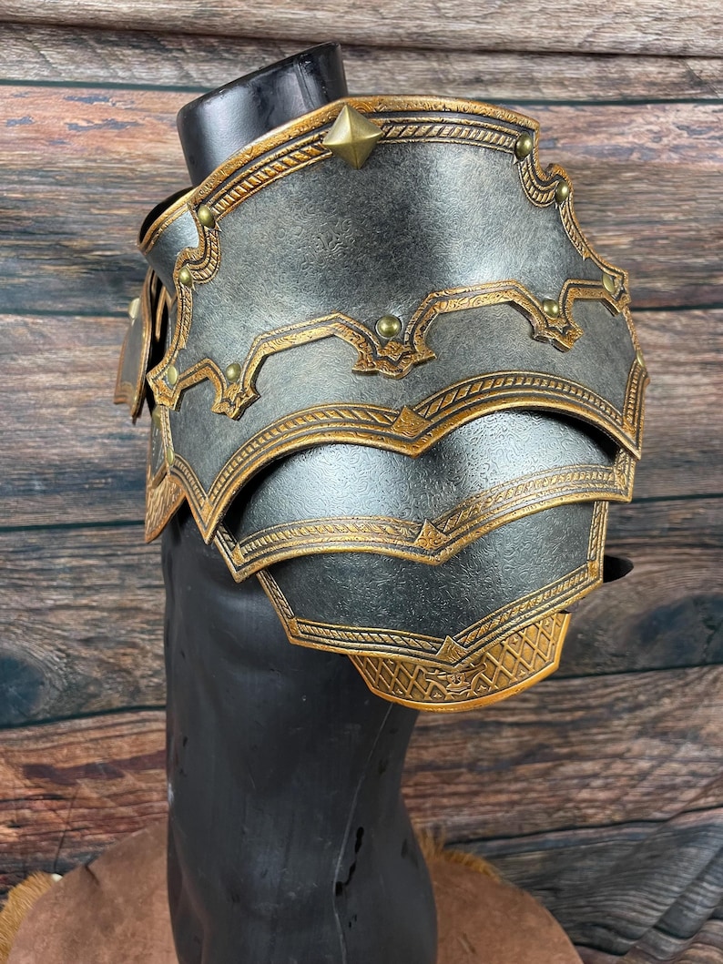 May include: A detailed, metallic shoulder armor piece in shades of gray and gold, displayed on a black mannequin. The armor features intricate embossed designs and decorative studs. The layered construction and ornate detailing suggest a fantasy or historical theme.