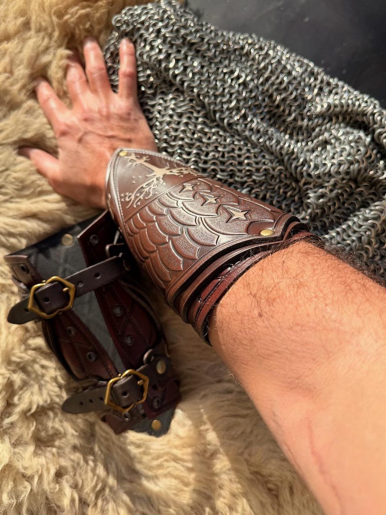 Puede incluir: Armadura de brazo de cuero marr&oacute;n con dise&ntilde;os de escamas y estrellas en relieve. La armadura se asegura con correas y hebillas de lat&oacute;n. Tambi&eacute;n se ve un protector de antebrazo de cuero marr&oacute;n a juego. El fondo incluye cota de malla y una superficie texturizada.