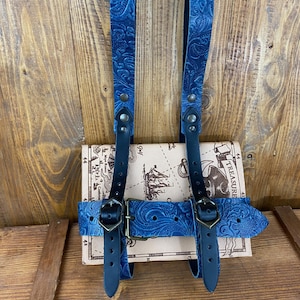 Puede incluir: Correas de cuero azul con un patrón en relieve floral, unidas a un libro con un diseño de mapa del tesoro pirata. Las correas tienen hebillas metálicas y están diseñadas para mantener el libro cerrado.