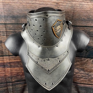 Può includere: Un gorgiera da cavaliere color argento, con un design rivettato e uno scudo decorativo con un emblema di teschio e corona d'alloro. L'armatura è esposta su un busto di manichino nero su sfondo di legno.
