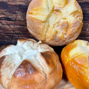 Puede incluir: Tres panes redondos y crujientes con una corteza dorada y una parte superior clara y texturizada. El pan se exhibe sobre una superficie de madera, mostrando los detalles y texturas de los productos horneados. Los panes varían en color, de marrón claro a oscuro.