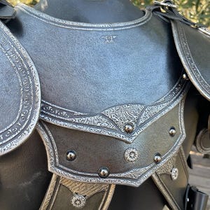 May include: Close-up of a medieval-style chest armor. The armor is primarily dark gray with intricate silver-colored decorative patterns and rivets. The shoulder pieces are detailed with embossed designs and leather straps.