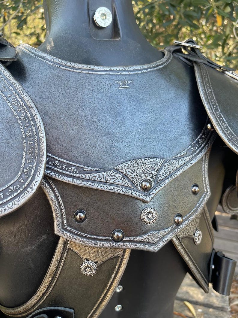 May include: Close-up of a medieval-style chest armor. The armor is primarily dark gray with intricate silver-colored decorative patterns and rivets. The shoulder pieces are detailed with embossed designs and leather straps.
