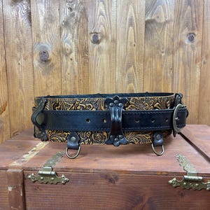 May include: A wide brown leather belt with a black leather strap and intricate floral tooling. The belt has three metal D-rings and a buckle with a decorative design.