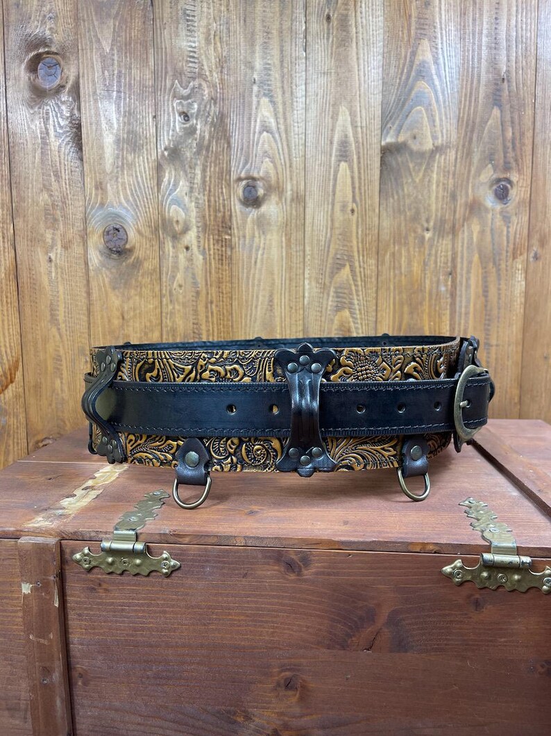 May include: A wide brown leather belt with a black leather strap and intricate floral tooling. The belt has three metal D-rings and a buckle with a decorative design.