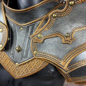 May include: Close-up of a black and gold shoulder armor piece. The armor features intricate embossed patterns, gold trim, and decorative rivets. The design suggests a medieval or fantasy theme.