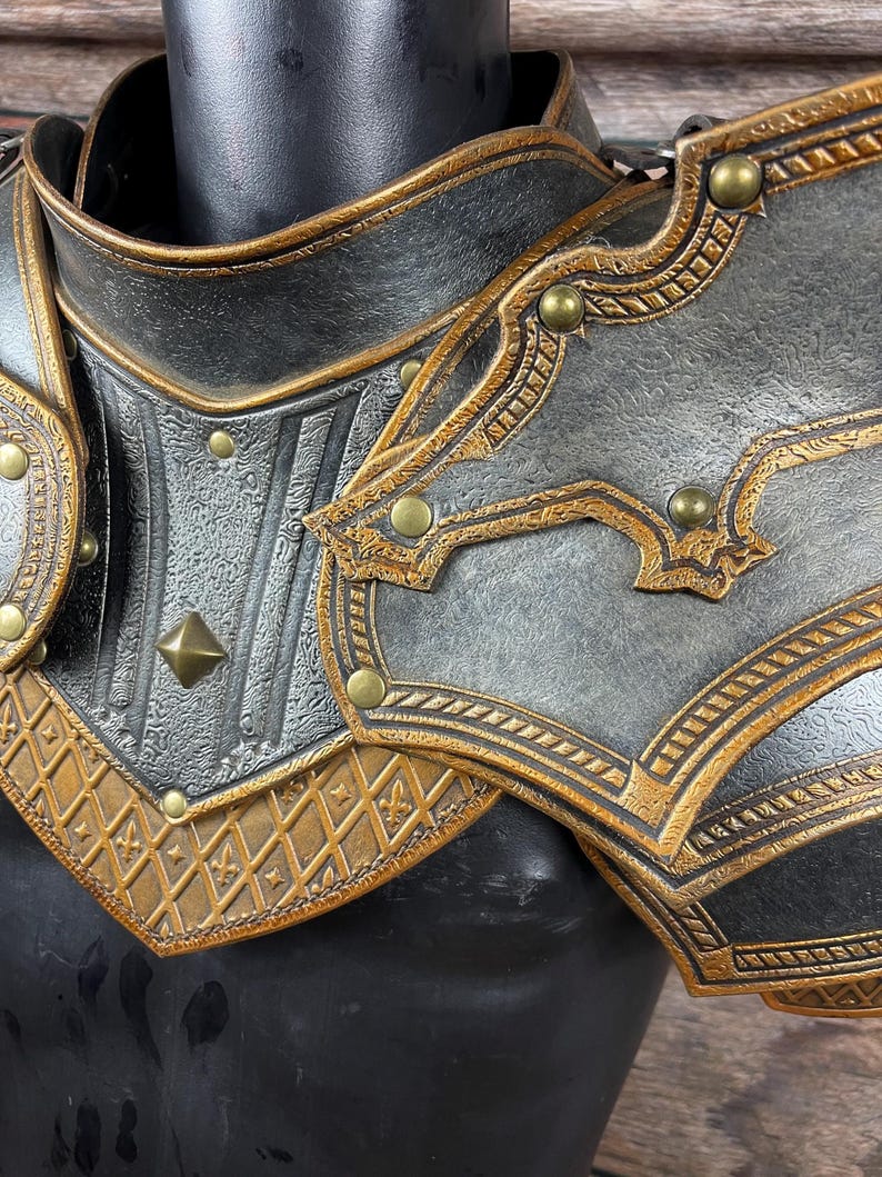 May include: Close-up of a black and gold shoulder armor piece. The armor features intricate embossed patterns, gold trim, and decorative rivets. The design suggests a medieval or fantasy theme.