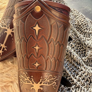 Puede incluir: Primer plano de dos protectores de brazo de cuero marr&oacute;n con dise&ntilde;os de estrellas y &aacute;rboles en relieve. Los protectores de brazo tienen un patr&oacute;n decorativo y est&aacute;n acentuados con detalles dorados. Una textura de cota de malla es visible en el fondo.