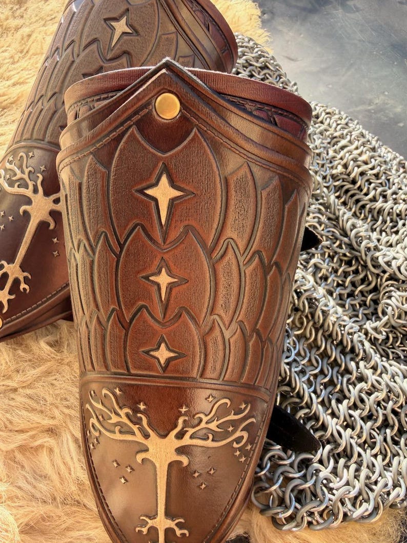 Puede incluir: Primer plano de dos protectores de brazo de cuero marr&oacute;n con dise&ntilde;os de estrellas y &aacute;rboles en relieve. Los protectores de brazo tienen un patr&oacute;n decorativo y est&aacute;n acentuados con detalles dorados. Una textura de cota de malla es visible en el fondo.