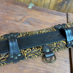 May include: A black leather belt with a gold embossed floral pattern. The belt has a metal buckle and a D-ring.