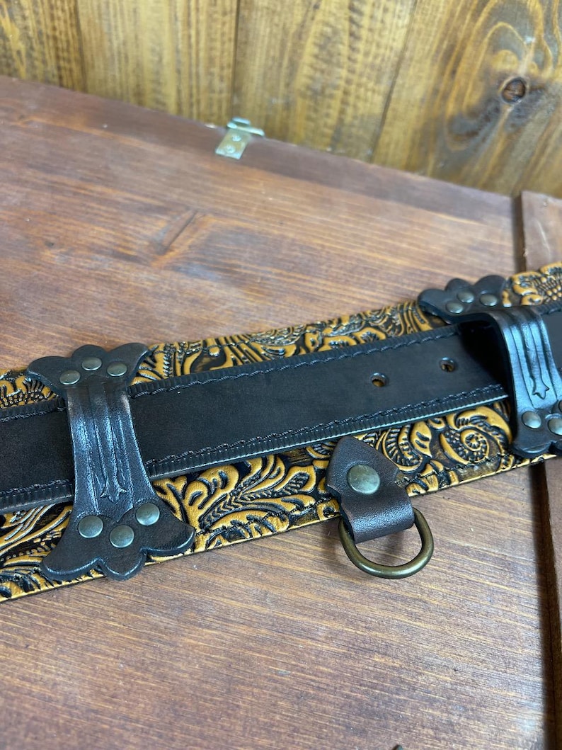 May include: A black leather belt with a gold embossed floral pattern. The belt has a metal buckle and a D-ring.