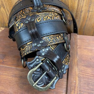 May include: A brown leather belt with a decorative buckle and multiple straps. The belt is adorned with intricate floral patterns and metal studs.
