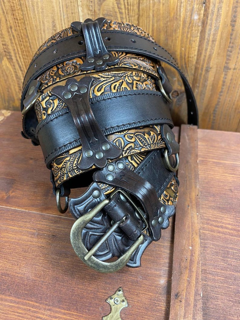 May include: A brown leather belt with a decorative buckle and multiple straps. The belt is adorned with intricate floral patterns and metal studs.