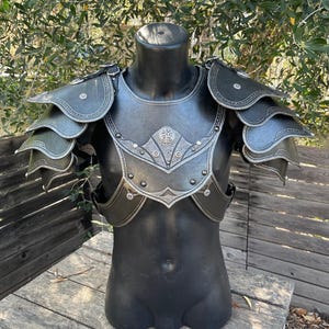 May include: Black and silver fantasy armor set displayed on a mannequin. The armor features layered shoulder pauldrons, a chest plate with decorative silver trim, and rivet details. The armor is designed to protect the torso and shoulders.
