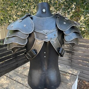 May include: Black and silver fantasy armor set displayed on a mannequin. The armor features layered shoulder pauldrons, decorative silver trim, and adjustable straps. The chest piece has a detailed design, suggesting a medieval or fantasy theme.