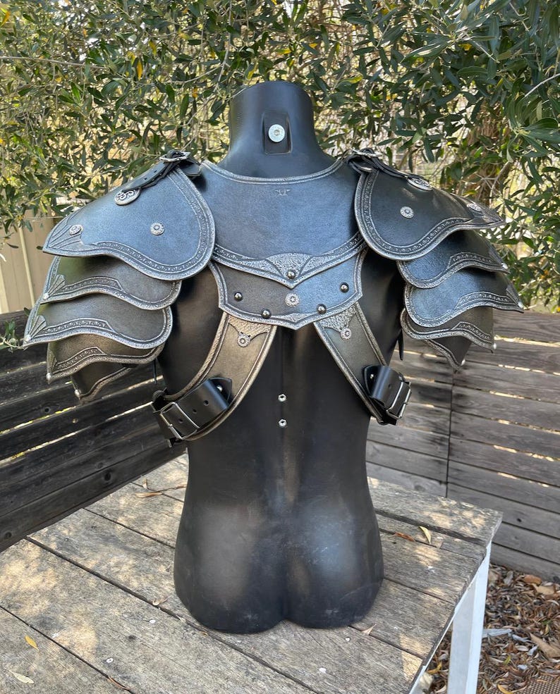 May include: Black and silver fantasy armor set displayed on a mannequin. The armor features layered shoulder pauldrons, decorative silver trim, and adjustable straps. The chest piece has a detailed design, suggesting a medieval or fantasy theme.