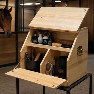 Peut inclure: Une boîte de pansage en bois clair avec un couvercle à charnière et un panneau avant rabattable. L'intérieur comporte deux étagères organisées avec divers accessoires de pansage, dont des vaporisateurs, des brosses, un cure-pied et un récipient noir.