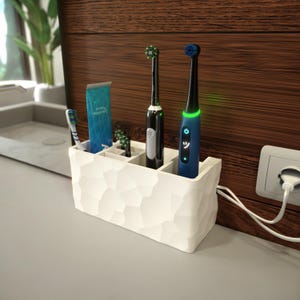 Könnte beinhalten: Weißer Zahnbürstenhalter mit vier Fächern, zwei elektrischen Zahnbürsten, einer Zahnpastatube und zwei manuellen Zahnbürsten. Die elektrischen Zahnbürsten sind an eine Steckdose angeschlossen.