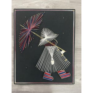 May include: String art piece depicting a person walking in the rain with an umbrella. The artwork features a black background with a frame. The person and umbrella are created with white, red, and blue string. The person is wearing red, white, and blue striped boots.