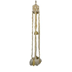 May include: A macrame plant hanger in cream and gold. The top section features a decorative design, with long, braided strands extending downwards. The bottom has a fringed edge. A bohemian style home decor item.