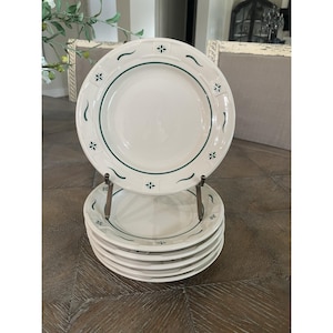 May include: A stack of six cream-colored ceramic plates featuring a dark green circular border and a rim decorated with alternating dark green floral motifs and curved line accents, displayed on a metal stand.