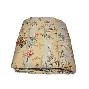 May include: Folded floral quilt featuring a pattern of roses and other flowers in shades of pink, blue, and cream. The quilt has a striped design and is folded neatly. A decorative bedding item.