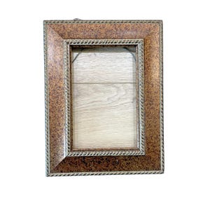 May include: Rectangular picture frame with a textured, bronze-colored finish. The frame has a rope-like border in a lighter gold tone. The interior opening reveals a light wood-grain backing. A small metal loop is attached for hanging.