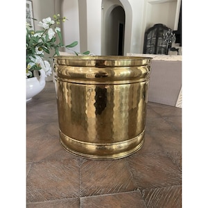 May include: A cylindrical brass planter featuring a hammered texture across its surface. The vessel has a smooth, reinforced rim at the top and a matching base, showcasing a polished metallic gold finish with subtle variations in light reflection.