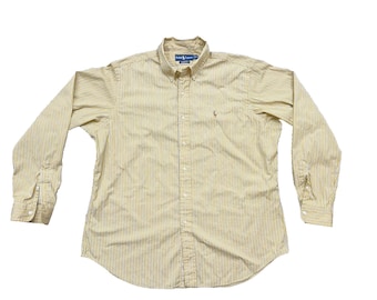 Pode incluir: Camisa de botão de manga comprida com padrão de riscas amarelas e brancas. A camisa tem gola abotoada, um pequeno logótipo bordado no peito e punhos abotoados. Feita de tecido leve.