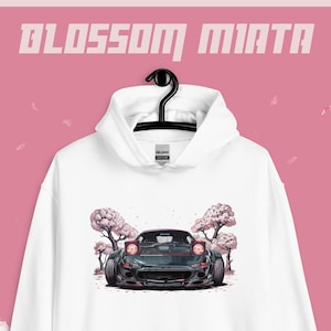 Könnte beinhalten: Weißer Kapuzenpullover mit einem Grafikdruck eines dunkelgrünen Autos mit rosa Kirschblütenbäumen. Der Text "BLOSSOM MIATA" ist oben aufgedruckt. Der Hoodie hat eine Vordertasche und eine Kapuze.