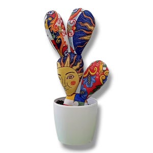 Könnte beinhalten: Ein dekorativer Stoffkaktus mit einem Sonnengesicht und bunten Mustern. Der Kaktus steht in einem weißen Topf. Der Stoff hat rote, blaue, gelbe und orange Farbtöne. Ein einzigartiges Wohnaccessoire.