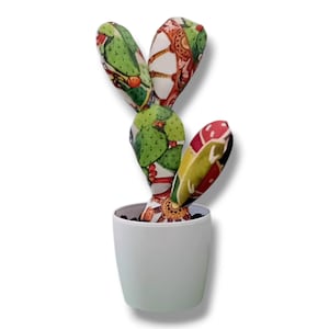 Könnte beinhalten: Ein Stoffkaktus in einem weißen Topf. Der Kaktus hat grüne Blätter mit einem Muster aus grünen Kakteen und weißen und roten floralen Designs. Der Topf ist weiß und steht auf einer weißen Oberfläche.