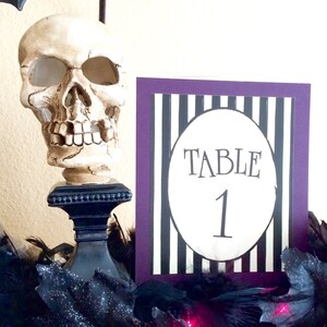 May include: A decorative Halloween table setting featuring a skull on a black stand, a table number card with black and white stripes, and the text "TABLE 1". The card has a purple border and is surrounded by black feathers.