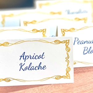 May include: White card with a gold decorative border and blue text that reads "Apricot Kolache".
