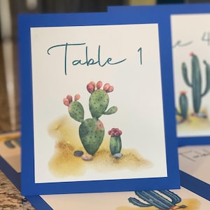 May include: A blue-bordered table card with a watercolor cactus illustration. The card reads "Table 1" in a script font. The cactus is green with pink accents, set against a sandy background.