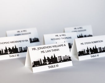 May include: White table cards with black text and a black silhouette of a city skyline. The text on the cards reads "Table 10" and includes the names of the guests.