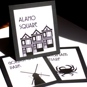 May include: Black and white art prints featuring iconic San Francisco landmarks. The prints include Alamo Square with Victorian houses, Golden Gate Park with a windmill, and Fisherman's Wharf with a crab illustration. Each print has a black border.