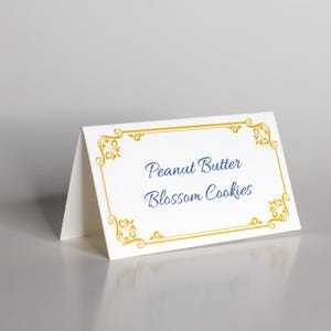 May include: White card with a gold border and a blue inscription that reads "Peanut Butter Blossom Cookies".