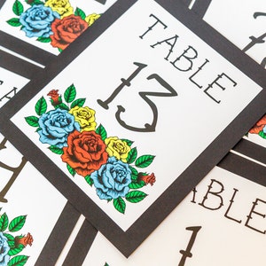 May include: White table number cards with black borders. Each card features the word "TABLE" and a number, with a floral design of red, yellow, and blue roses. The cards are arranged at an angle.