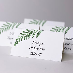 Puede incluir: Una tarjeta de sitio plegada de color blanco con una ilustración de hoja de helecho verde impresa en la esquina superior izquierda. La tarjeta muestra el texto impreso en negro Niecy Johnson y Table 15 en una fuente cursiva y sans-serif.